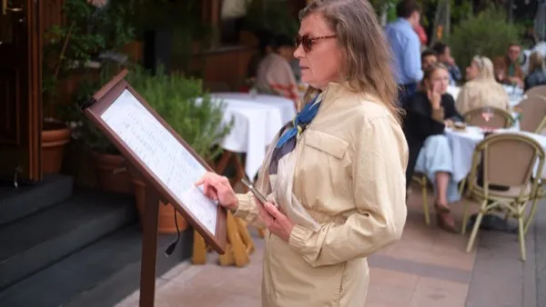 A woman reading a printed menu stand on the pavement outside a street cafe.