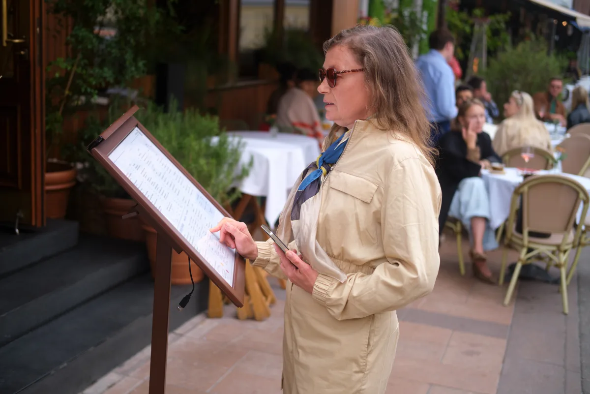 A woman reading a printed menu stand on the pavement outside a street cafe.