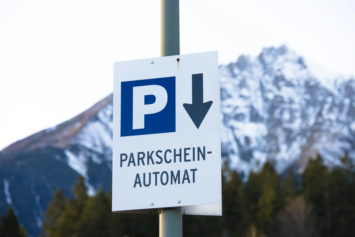 A German Parkscheinautomat parking sign on a pole against snow-covered mountains.